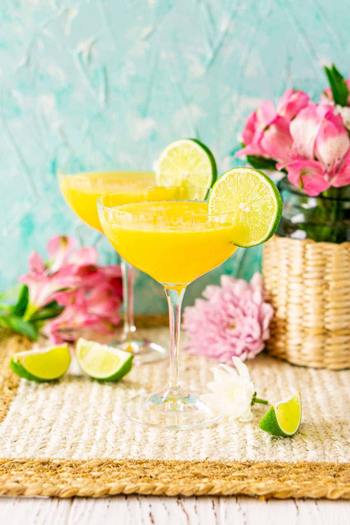 A passion fruit daiquiri on a straw placemat against a blue background with limes and colorful flowers around it.