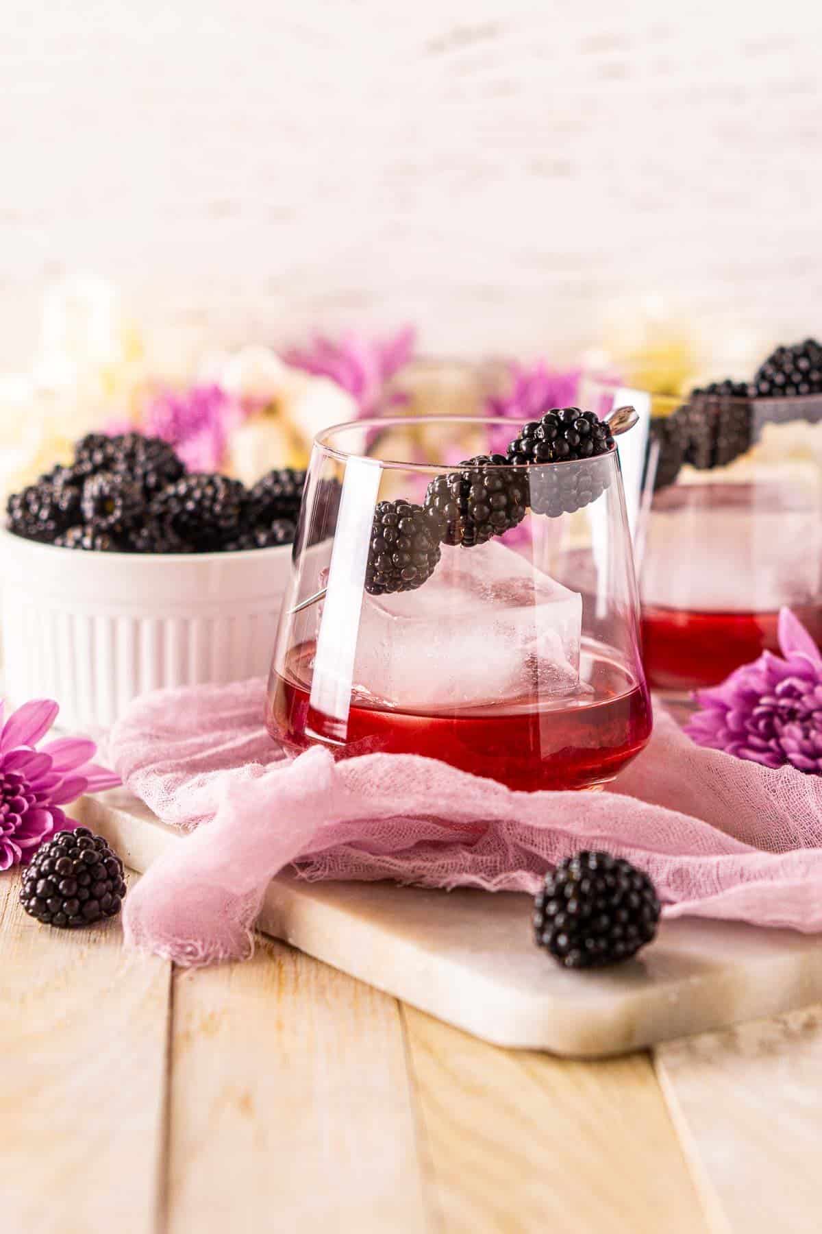 A blackberry old fashioned on a lightly colored wooden board with purple flowers to the side.