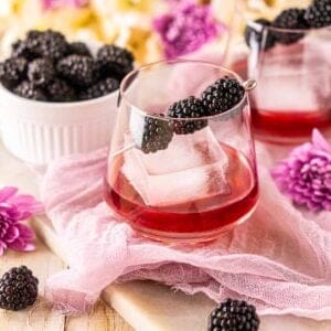 A blackberry old fashioned on purple cloth with fresh berries and flowers surrounding it.