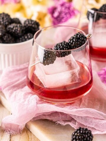 A blackberry old fashioned on purple cloth with fresh berries and flowers surrounding it.