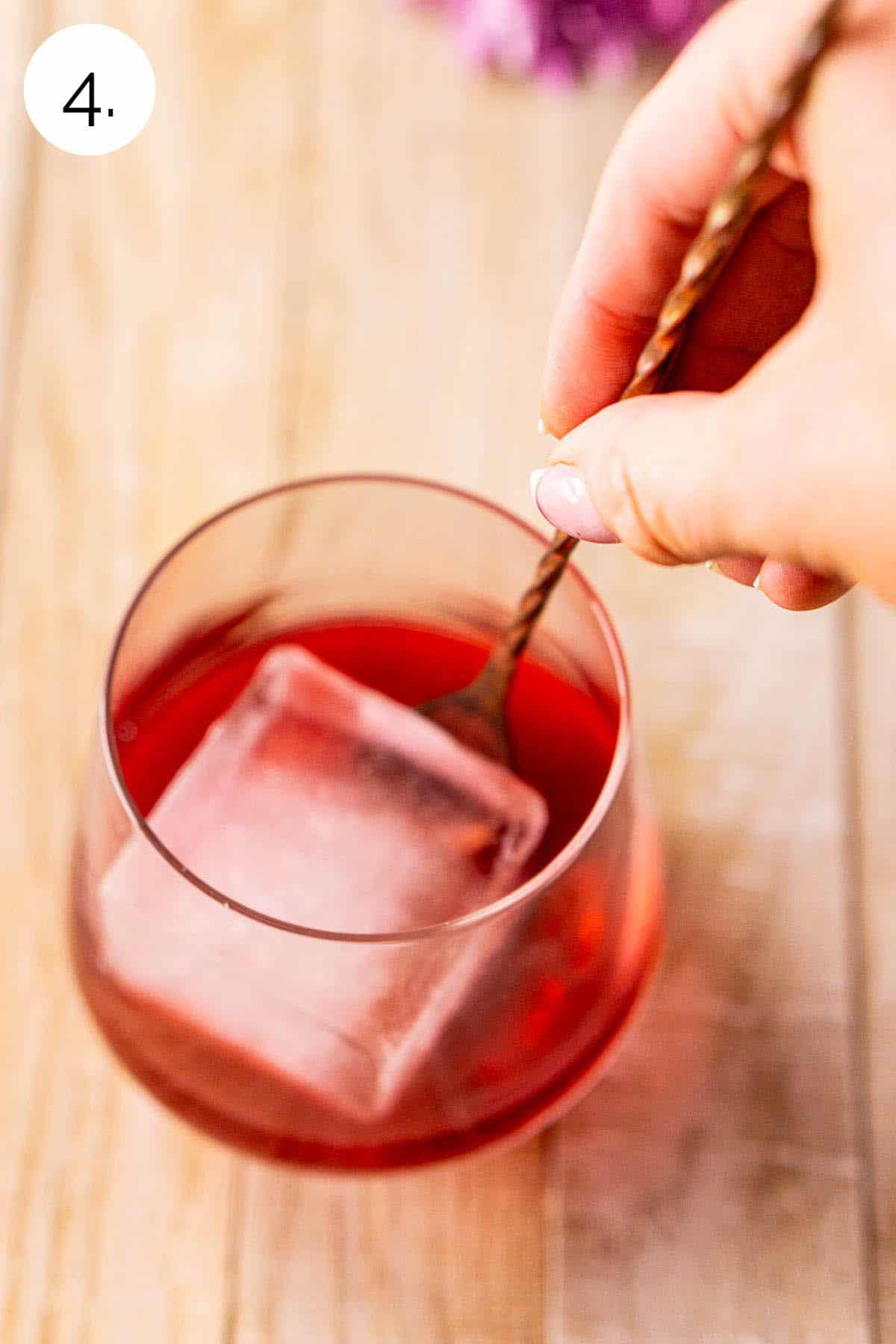 A hand stirring the blackberry old fashioned with one large ice cube to mix the drink on a cream-colored wooden surface.