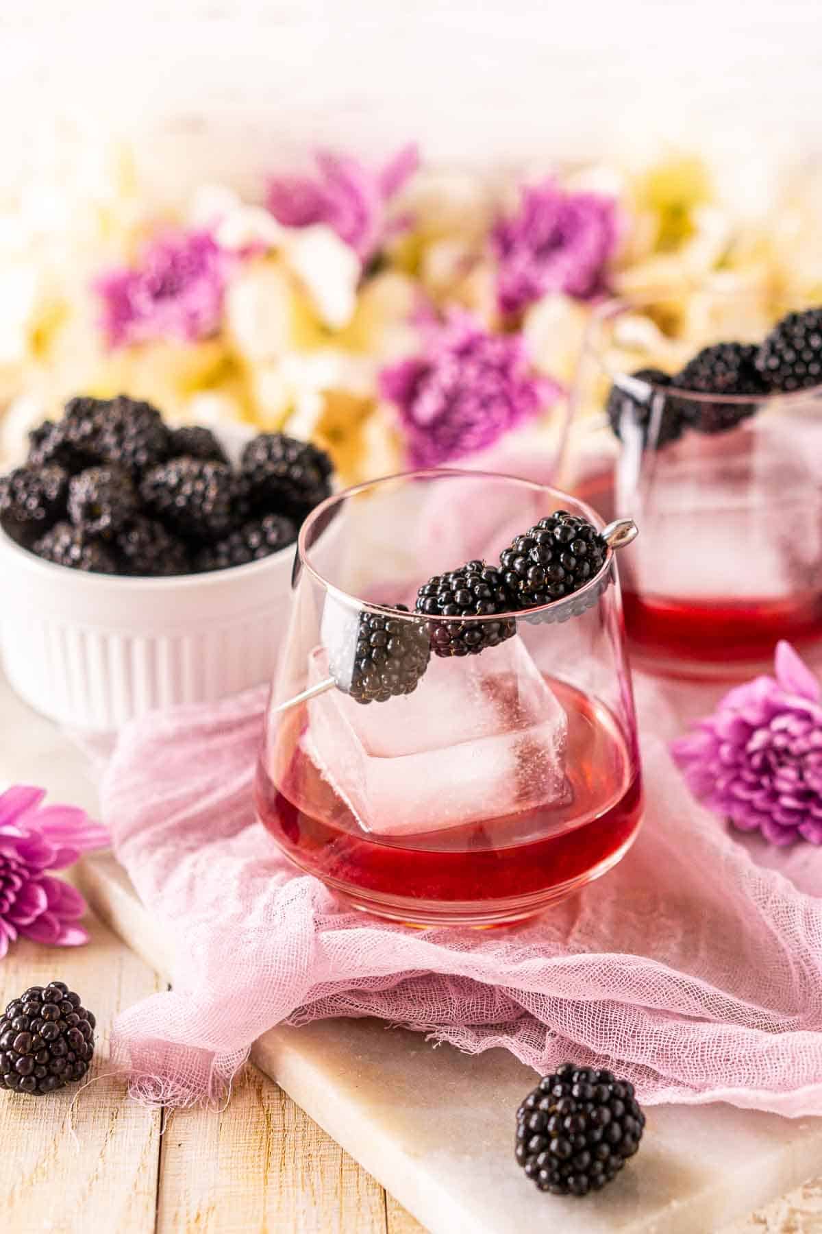 A blackberry old fashioned on purple cloth with fresh berries and flowers surrounding it.