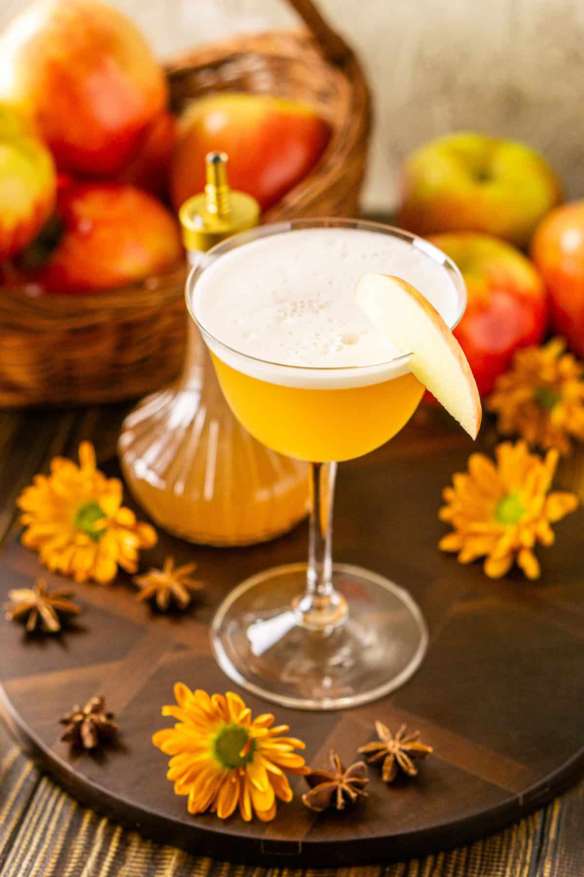 A side angle of the apple cider bourbon sour on a wooden serving tray with star anise and fall flowers scattered around it.