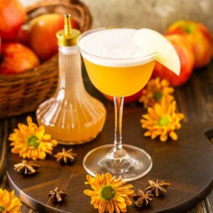 An apple cider whiskey sour on a brown serving tray with apples in the background and spices and yellow fall flowers around it.