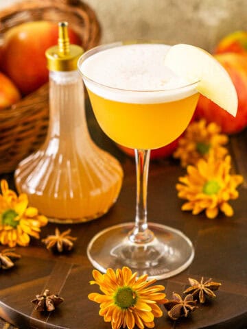 An apple cider whiskey sour on a brown serving tray with apples in the background and spices and yellow fall flowers around it.