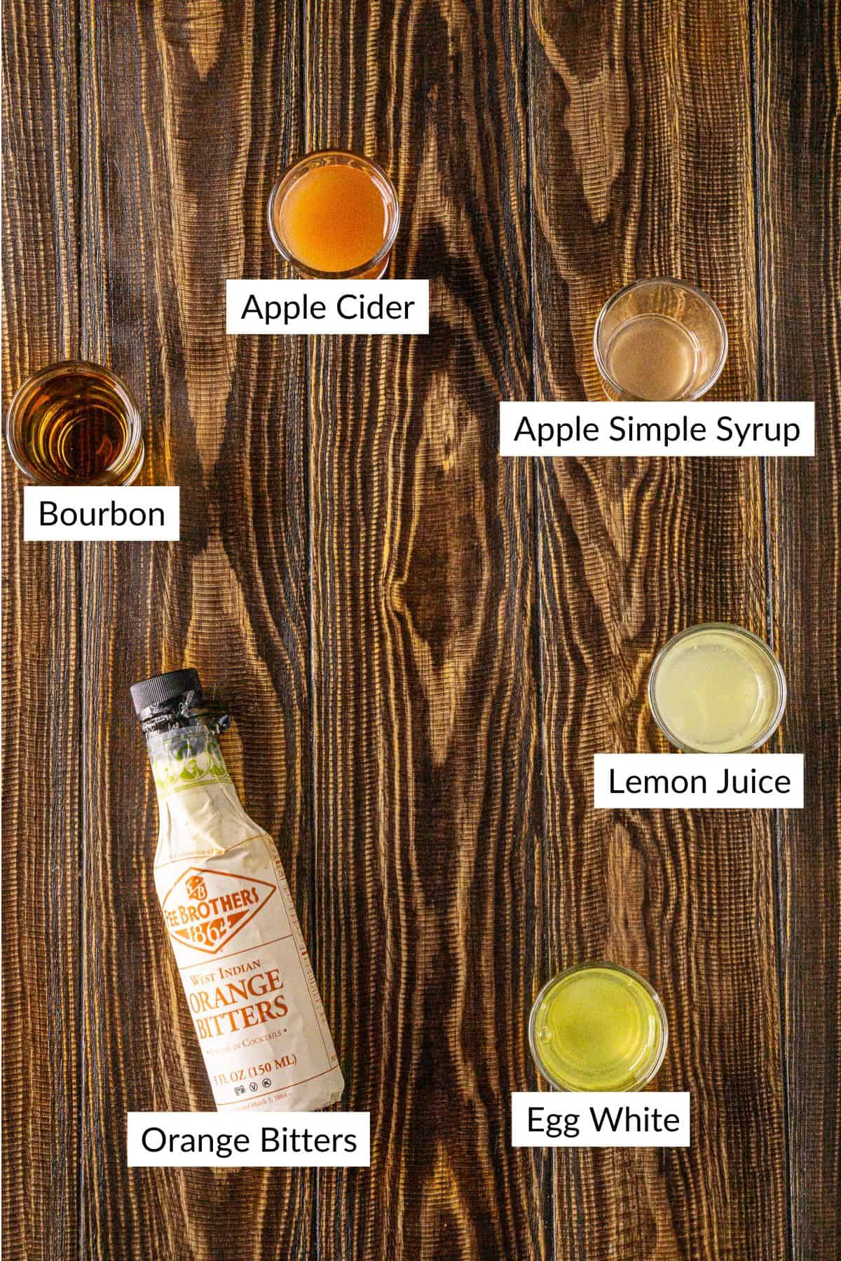 The apple cider whiskey sour ingredients on a brown wooden board with white and black labels by the items.