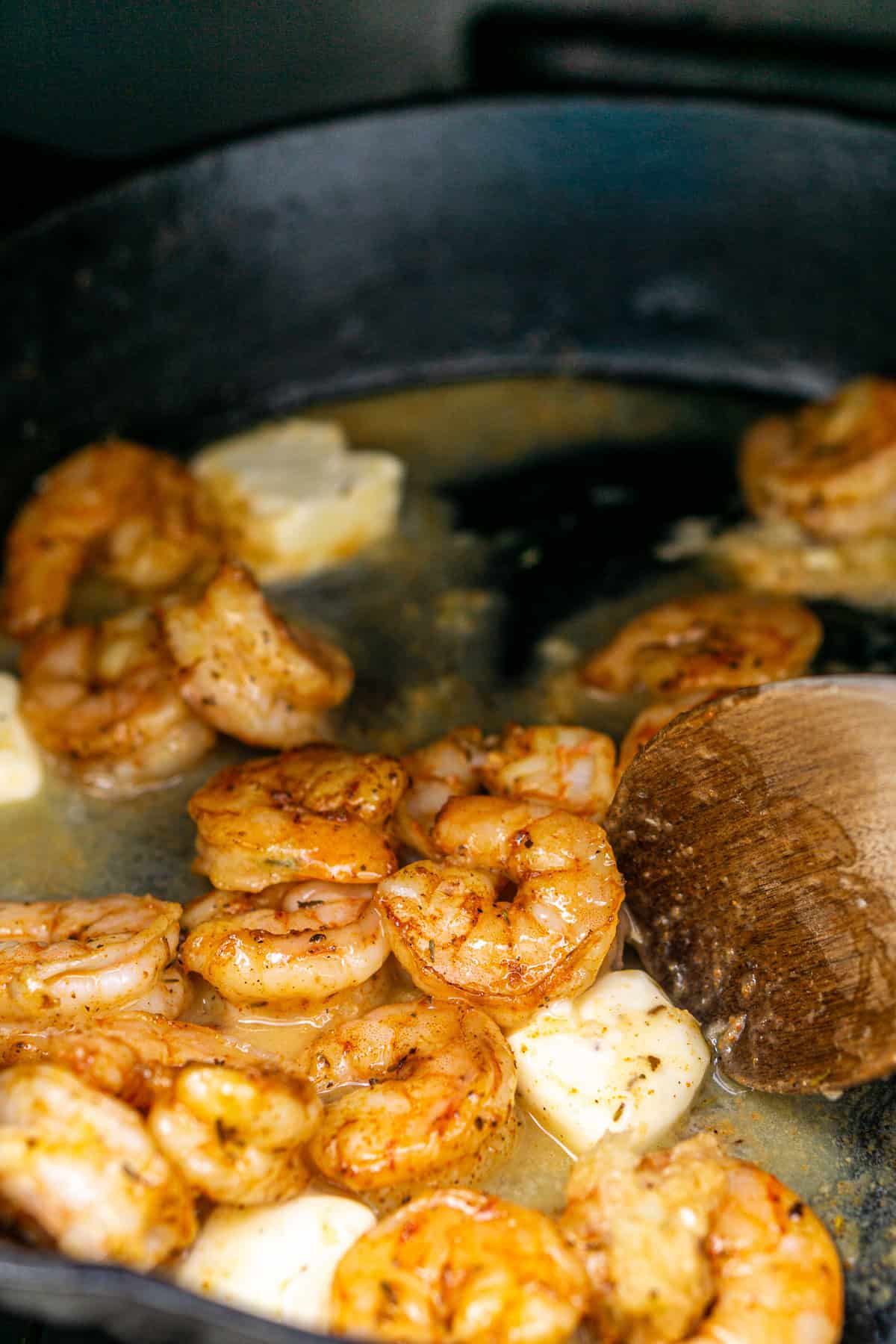 A wooden spoon stirring the butter and garlic in a black cast-iron skillet with the shrimp.