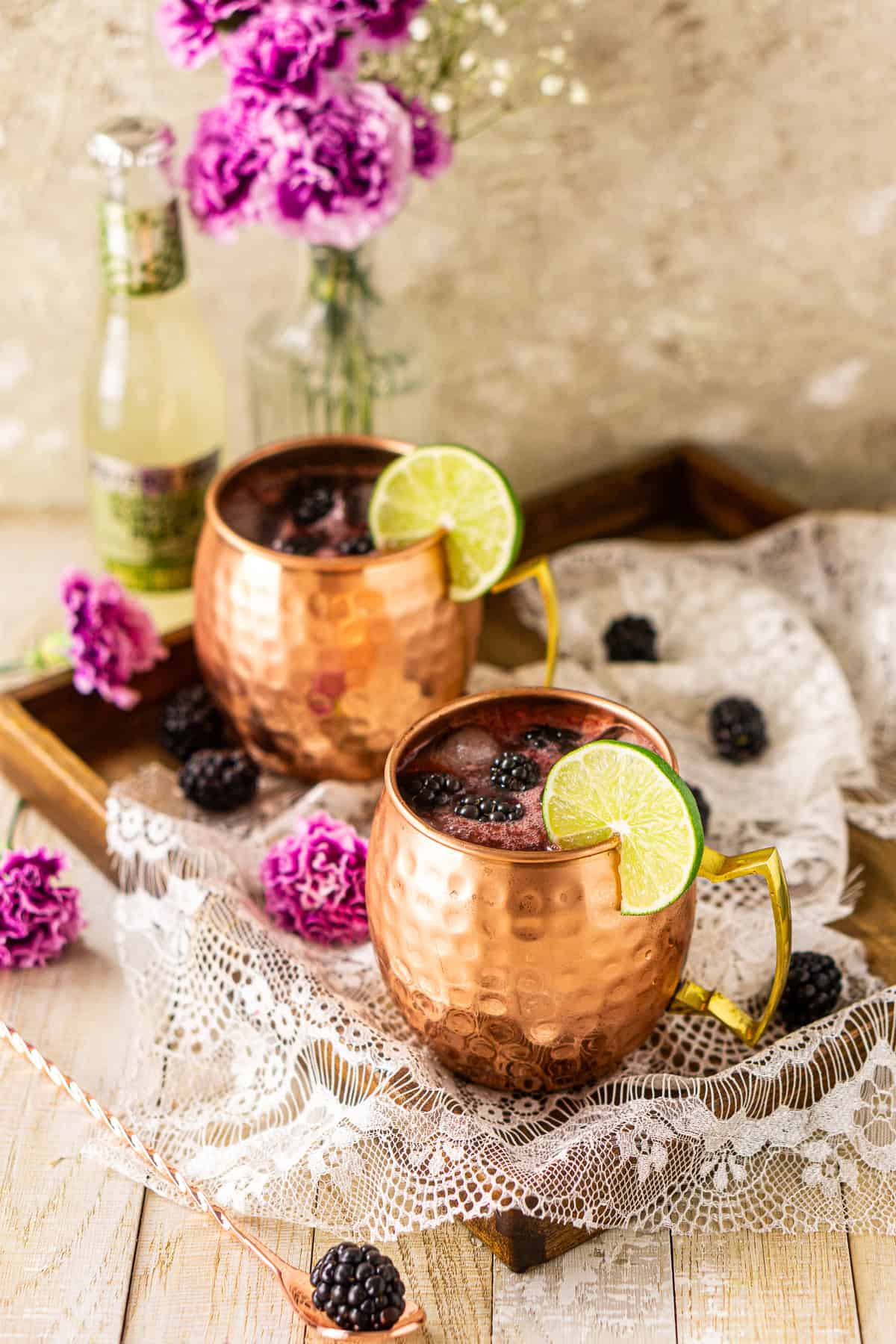 Two blackberry mules on a wooden tray covered in white lace with purple flowers and berries scattered around them.