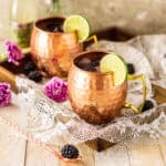 A blackberry Moscow mule on a wooden tray with a copper bar spoon and a fresh berry to the left and a bottle of ginger beer behind it.