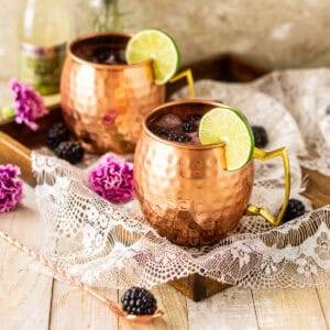 A blackberry Moscow mule on a wooden tray with a copper bar spoon and a fresh berry to the left and a bottle of ginger beer behind it.