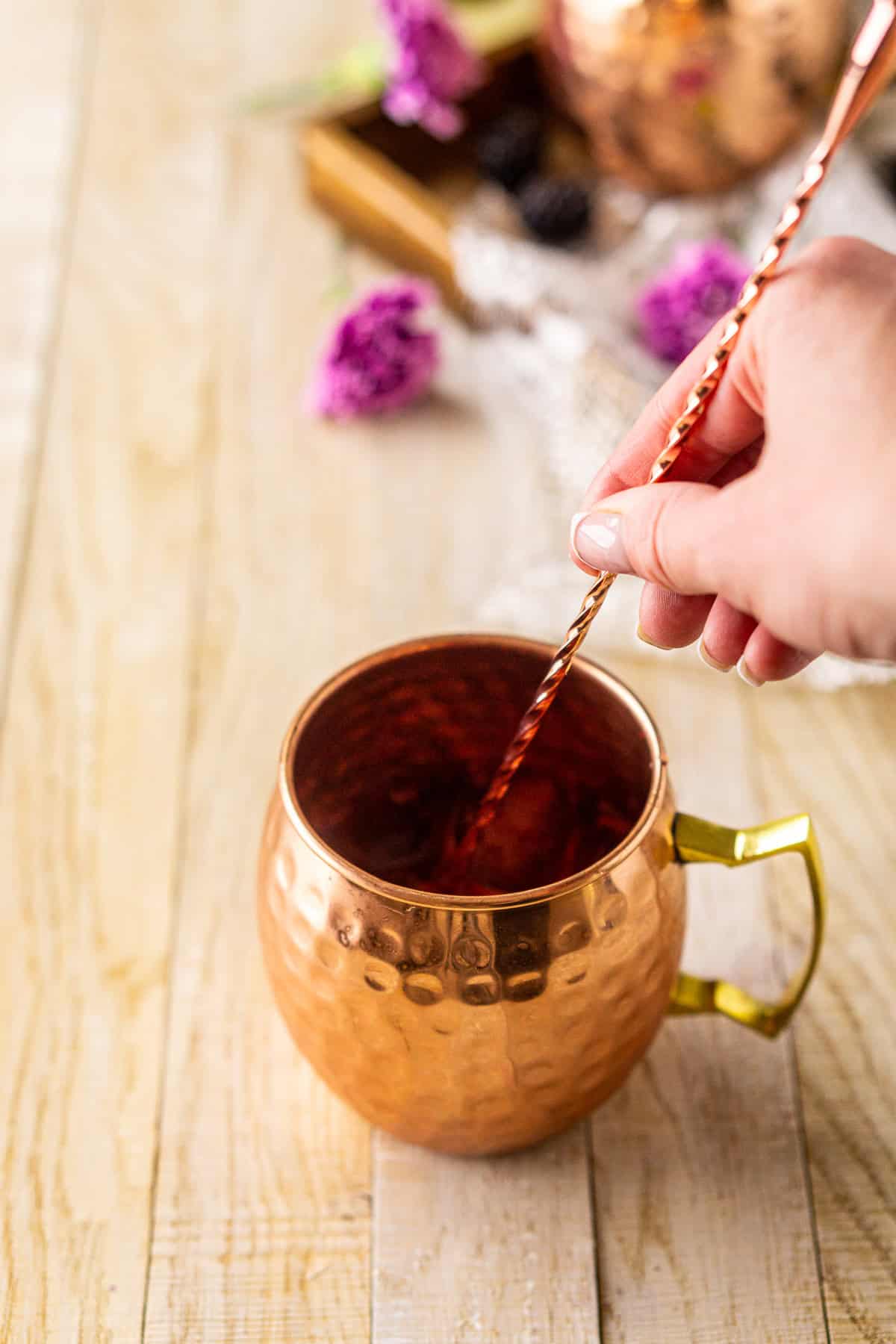A hand stirring the blackberry mule with a copper bar spoon on a cream-colored wooden surface to mix the drink.