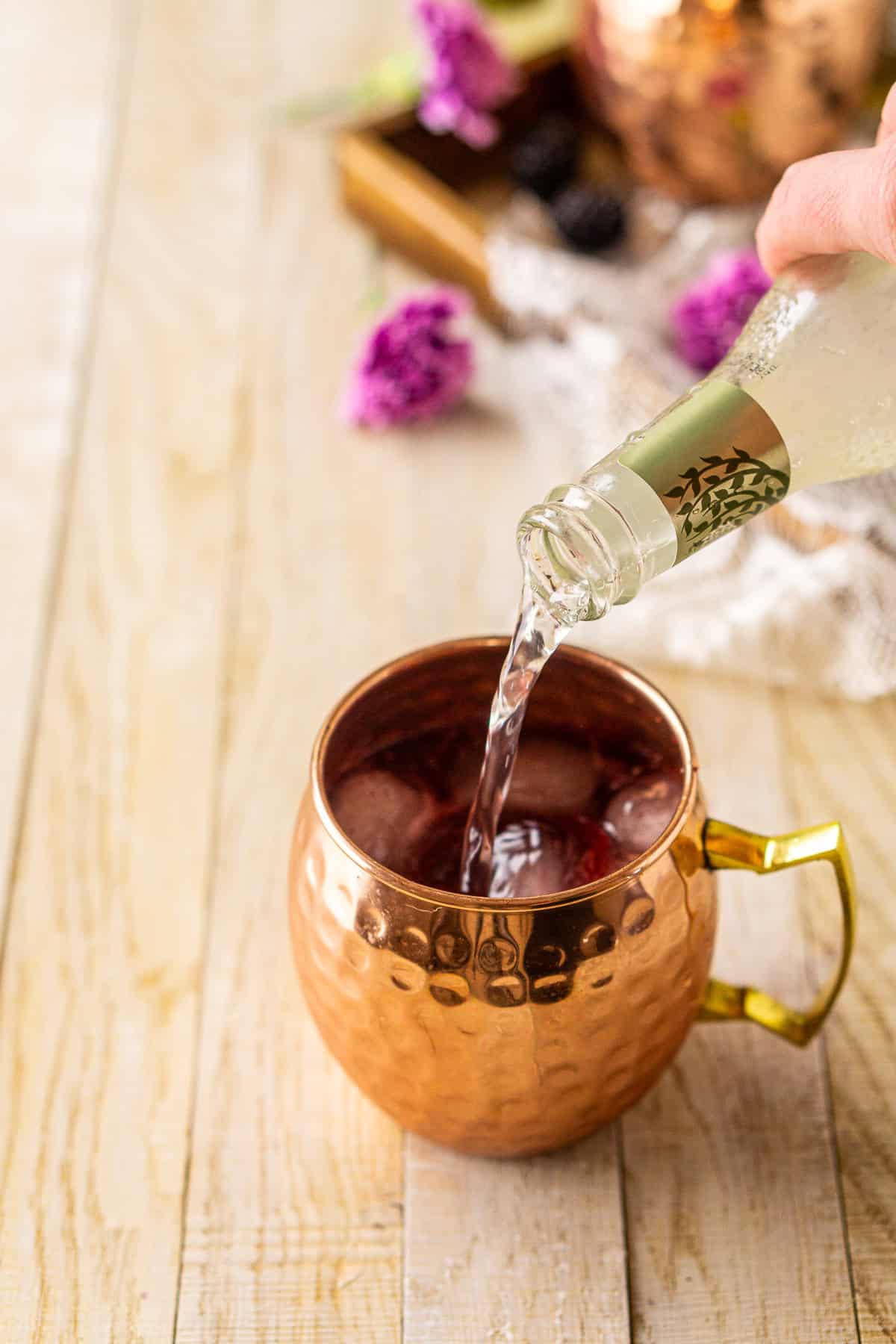 A hand pouring a bottle of ginger beer into the copper mug with the blackberry mixture on a cream-colored wooden surface.