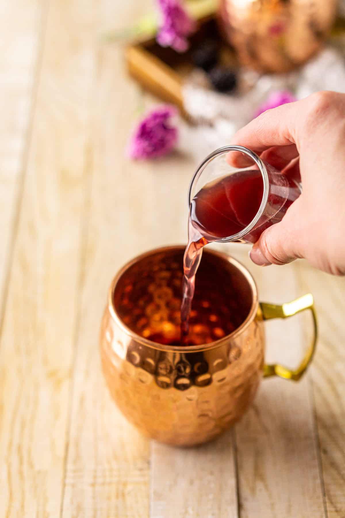 A hand pouring a shot glass of blackberry vodka into a copper mug on a cream-colored wooden surface.