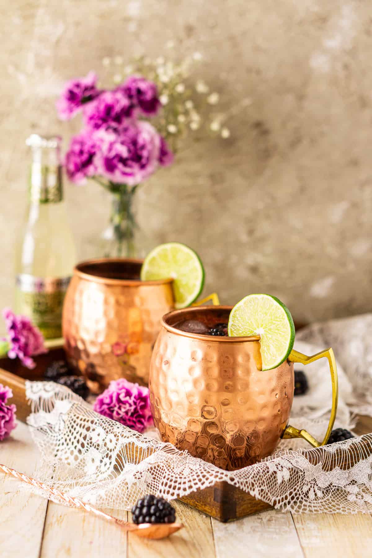 A blackberry Moscow mule on a wooden tray with a bottle of ginger beer and purple flowers in the background.