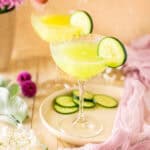 A cucumber martini on a plate with a purple cloth on the right and flowers in the background.