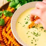 A hand dipping a sweet potato fry into a bowl of mango-habanero aioli with more fries and a bundle of cilantro on the side.