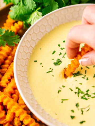 A hand dipping a sweet potato fry into a bowl of mango-habanero aioli with more fries and a bundle of cilantro on the side.