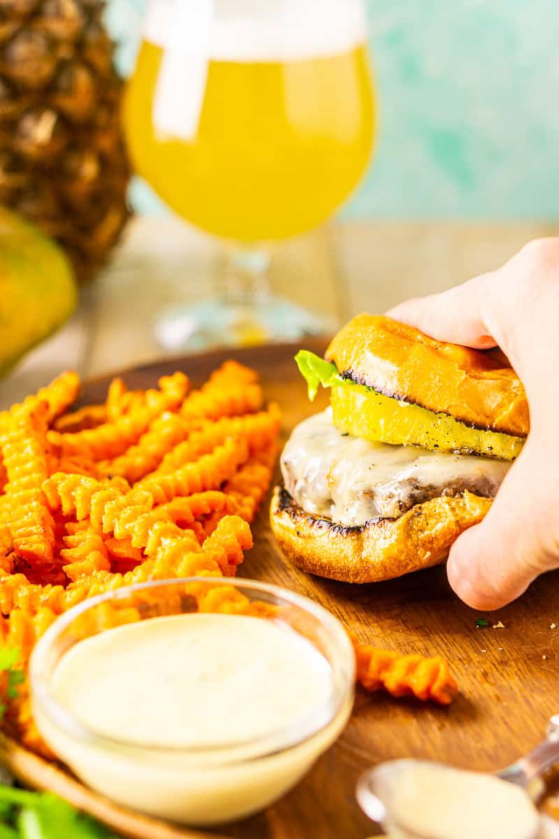 A hand grabbing a jerk burger off a brown wooden plate with sweet potato fries and mango aioli to the side.