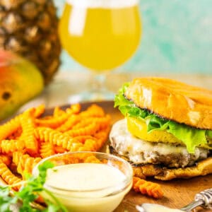 A jerk burger and bowl of mango-habanero aioli on a wooden plate with a beer and pineapple in the background.