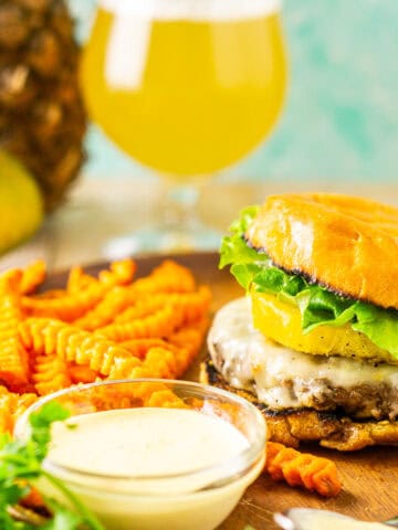 A jerk burger and bowl of mango-habanero aioli on a wooden plate with a beer and pineapple in the background.