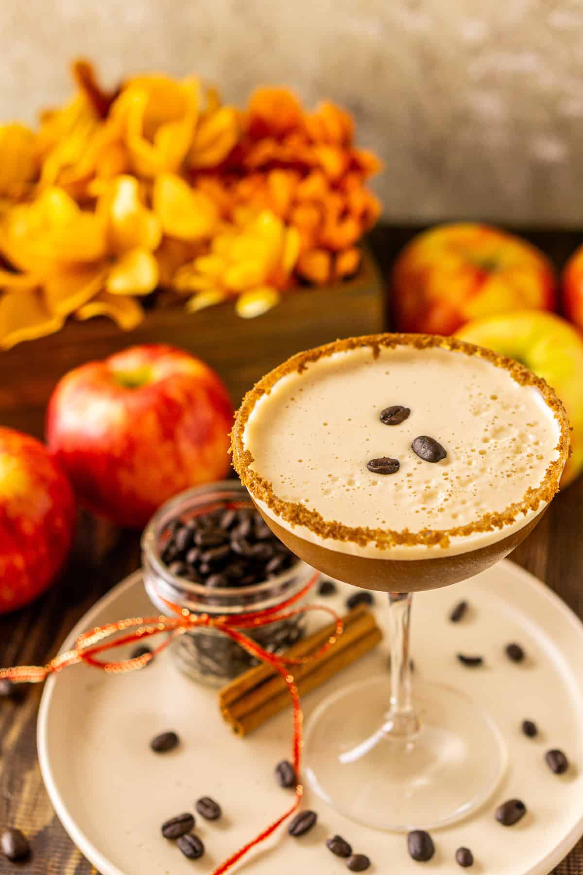 An apple espresso martini with coffee beans scattered around it and several apples in the background on a wooden surface.