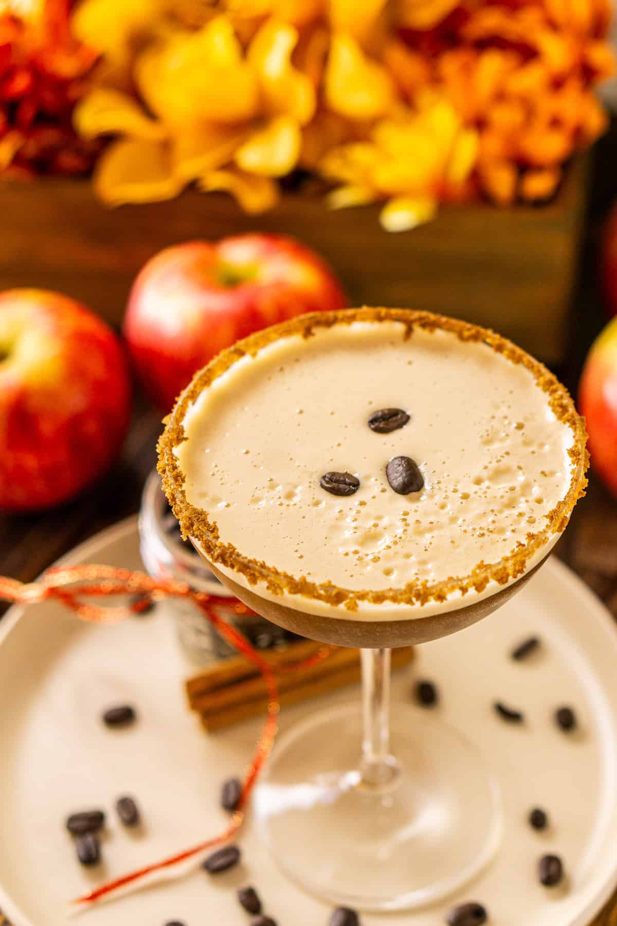 A close-up overhead shot of an apple espresso martini with three coffee beans placed on the drink's foam with fall flowers behind it.