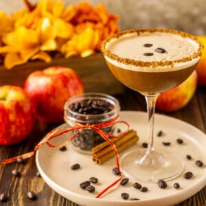 A spiced apple espresso martini on a cream-colored plate with coffee beans around it and fall flowers in the background.