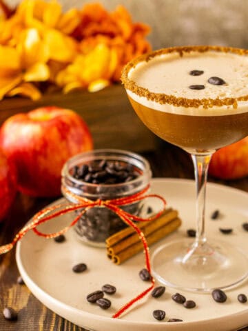 A spiced apple espresso martini on a cream-colored plate with coffee beans around it and fall flowers in the background.