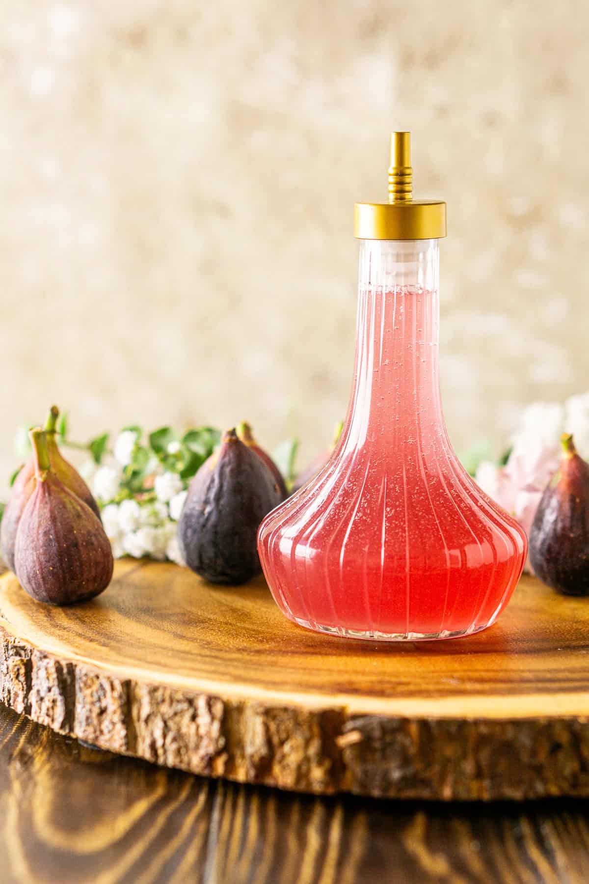 A bottle of fig simple syrup against a cream-colored background on a wooden serving platter with more figs behind it.