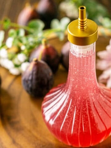 A bottle of fig simple syrup on a wooden serving platter with flowers and fresh figs scattered in the background.