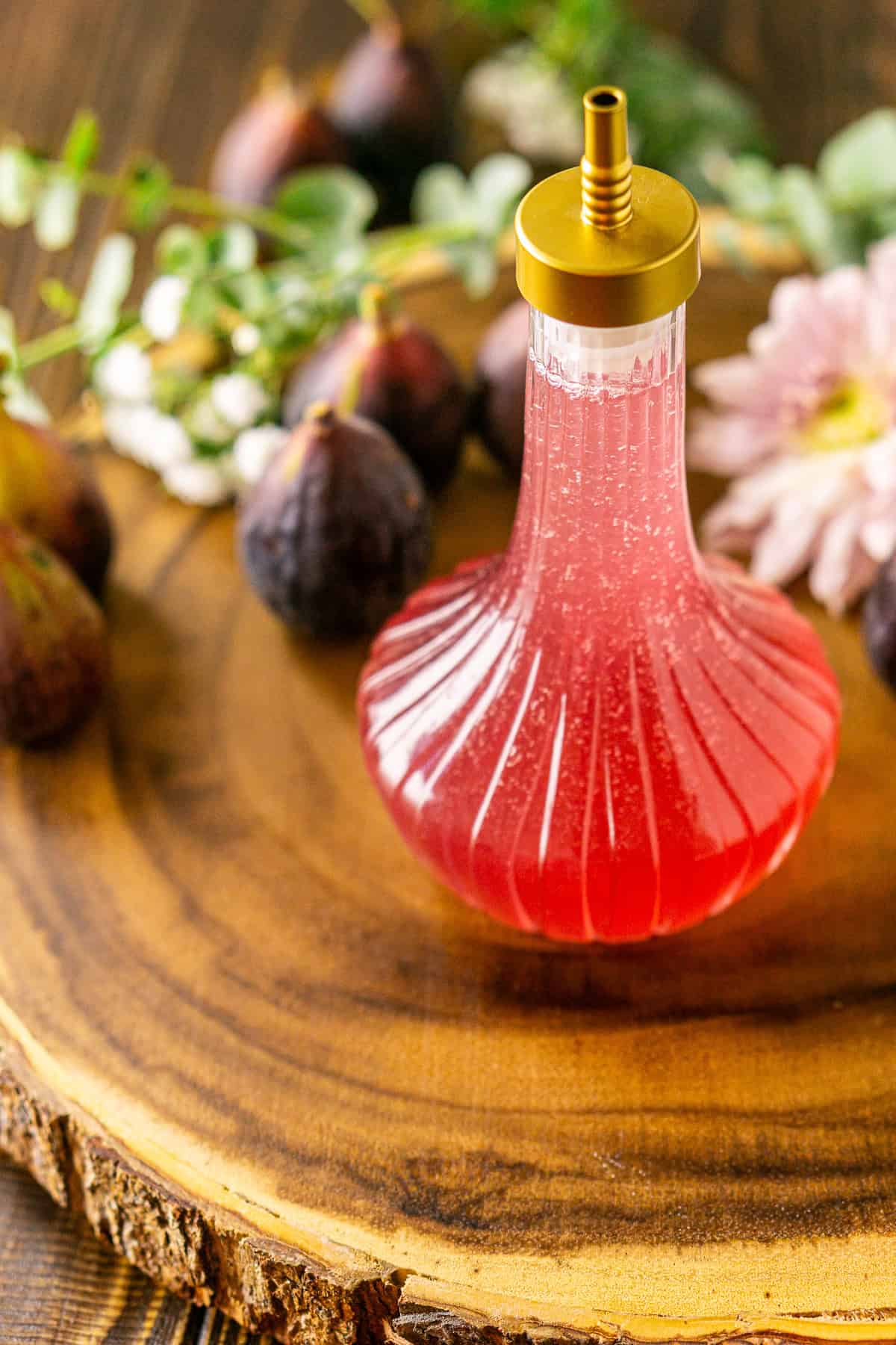 A bottle of fig simple syrup on a wooden serving platter with flowers and fresh figs scattered in the background.