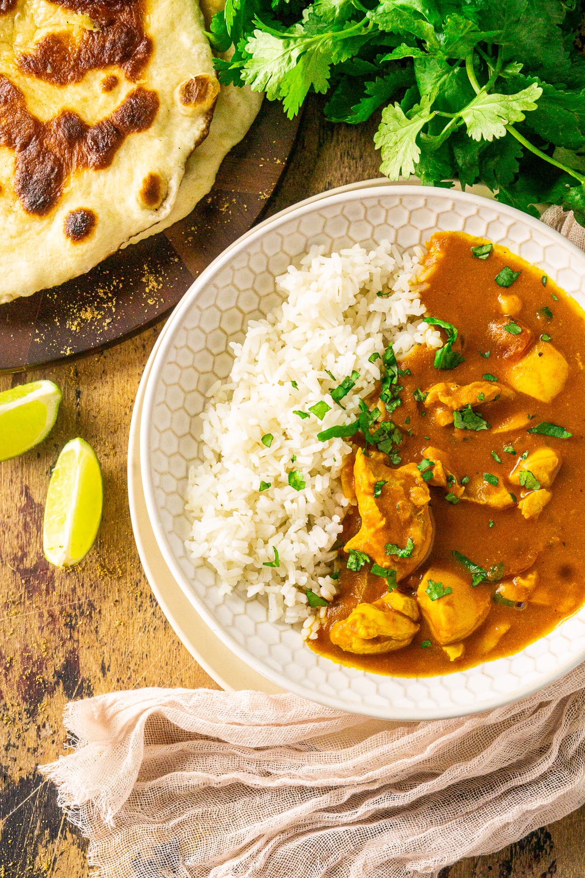 An aerial view of a bowl of pumpkin and chicken curry with a plate of naan and fresh cilantro in the top corners.