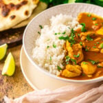 A bowl of pumpkin and chicken curry on a wooden surface with two lime slices to the side and naan and cilantro in the background.