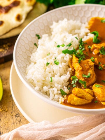 A bowl of pumpkin and chicken curry on a wooden surface with two lime slices to the side and naan and cilantro in the background.