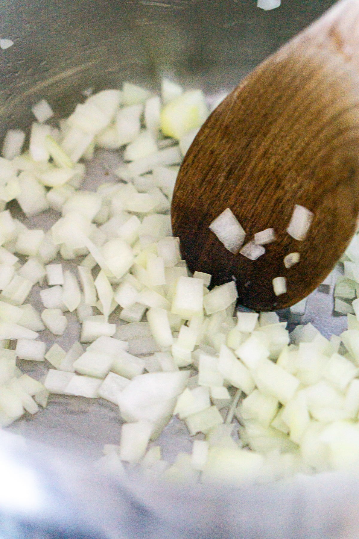 A brown wooden spoon cooking onions until they're softened in a stainless-steel pot.