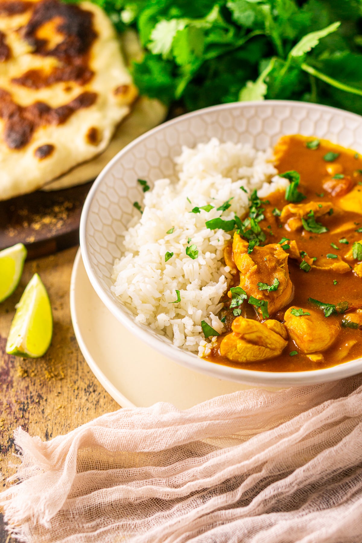 A bowl of pumpkin and chicken curry on a wooden surface with two lime slices to the side and naan and cilantro in the background.